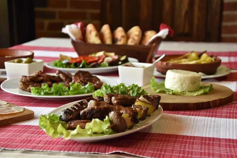 Plates of grilled qebapa with salad on a red checked tablecloth, bread behind