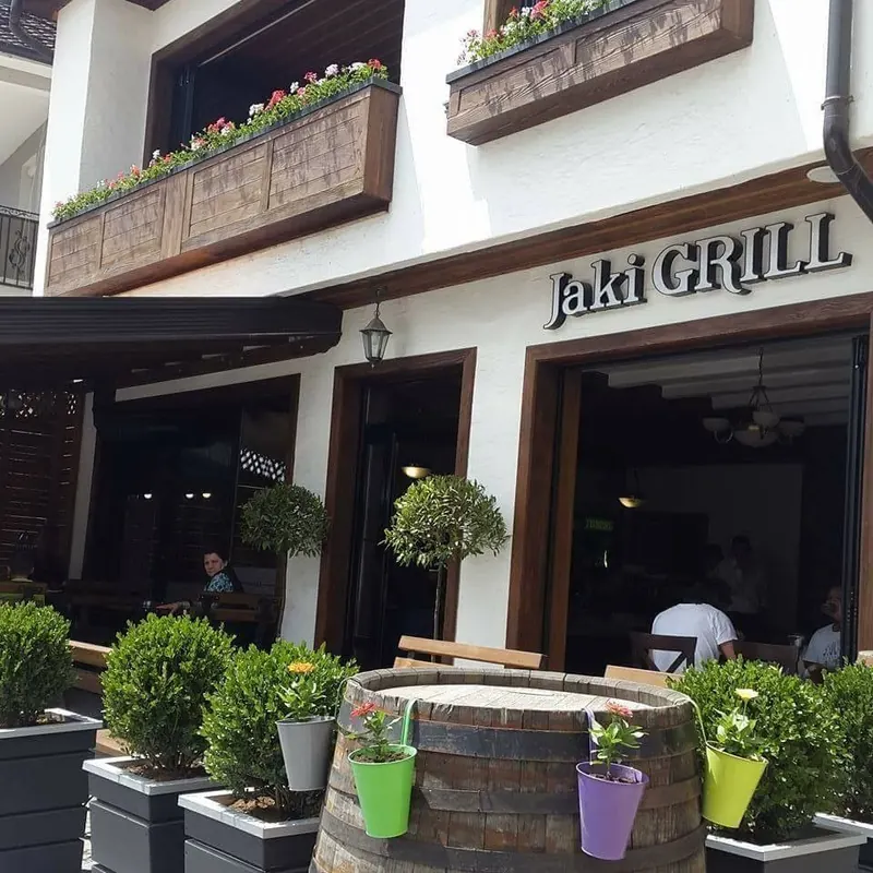 The white façade of Jaki Grill with its sign, flower boxes and a barrel of flowers by the door